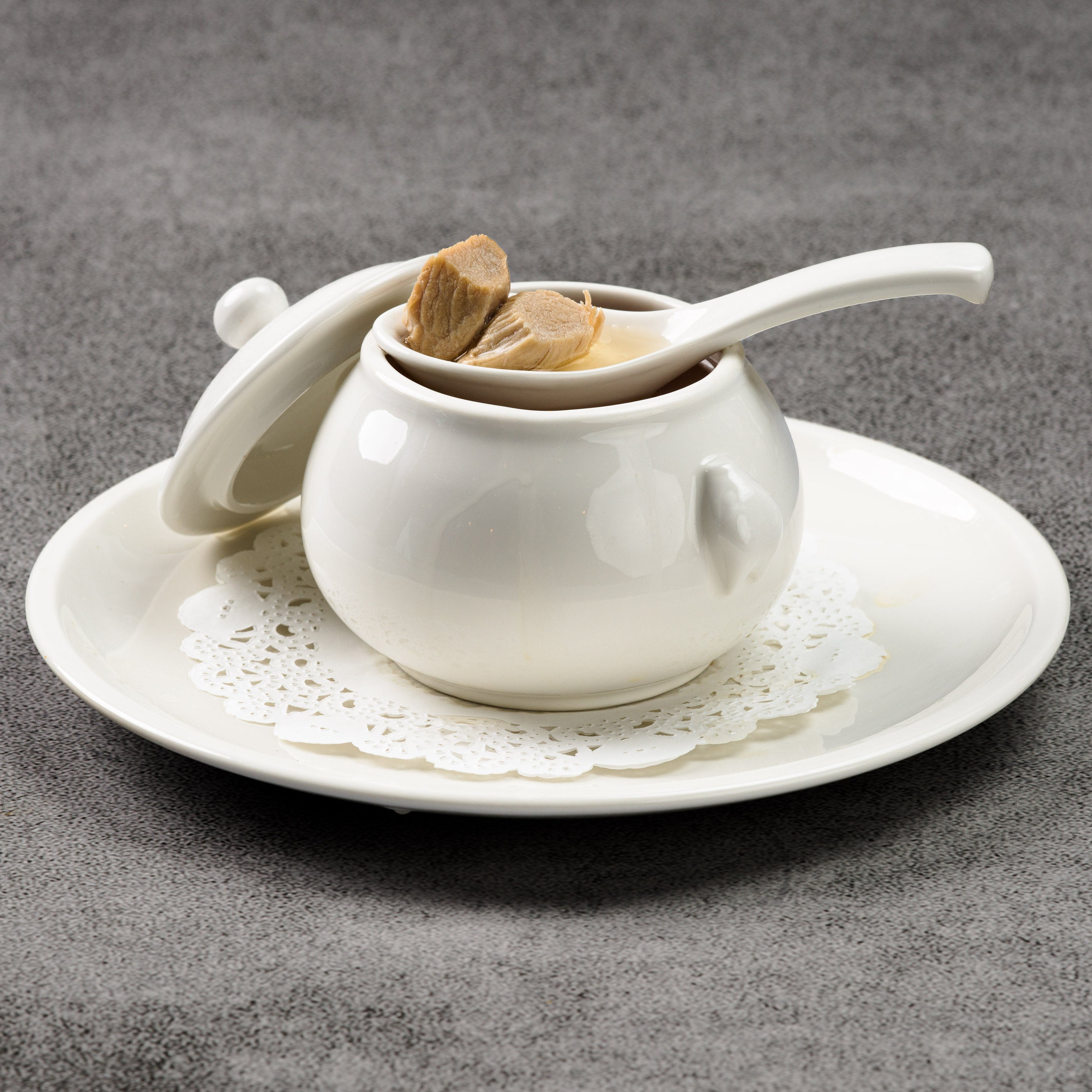White ceramic teacup of chicken soup with lid and spoon on a gray surface
