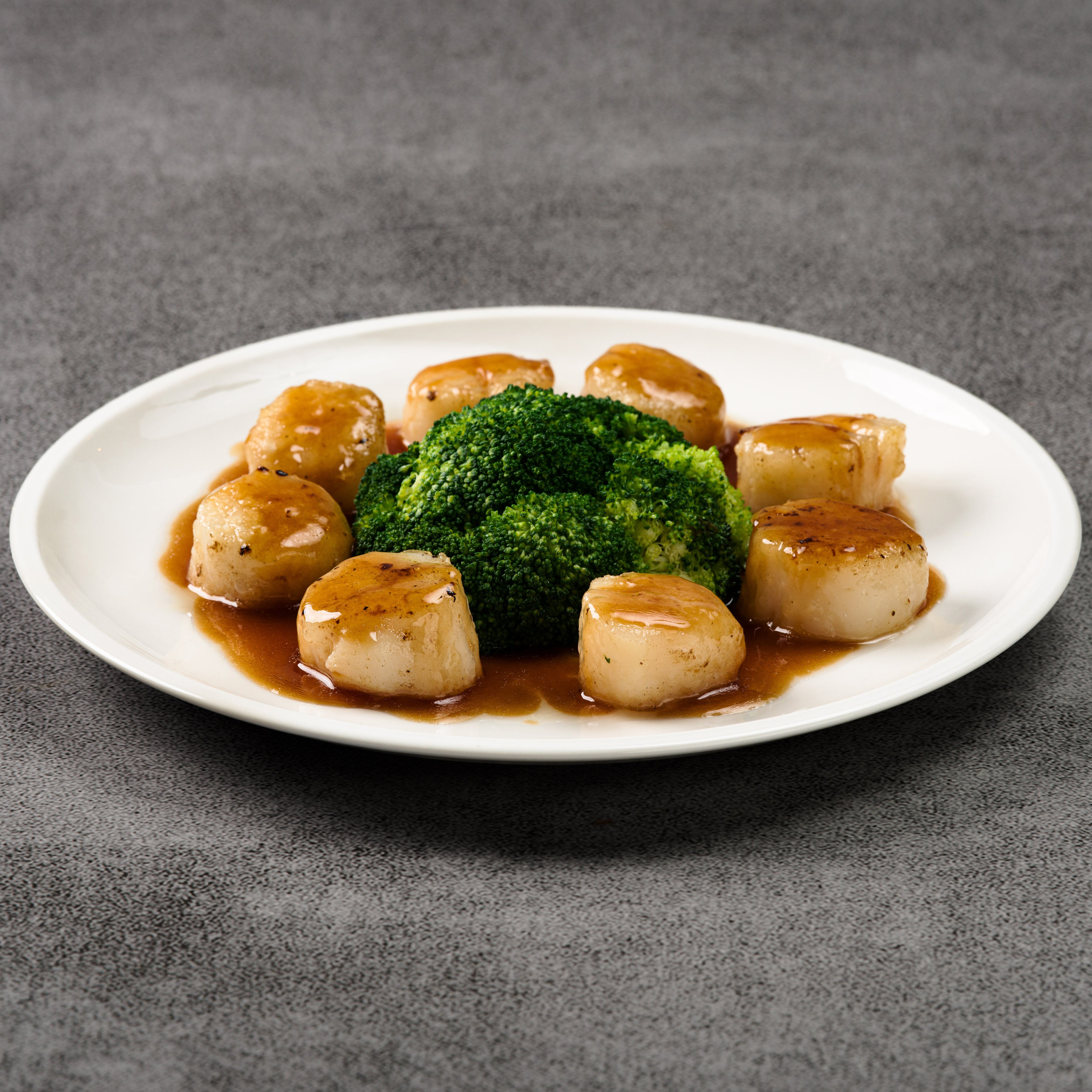 White plate with scallops and broccoli with a brown soy sauce on a gray background