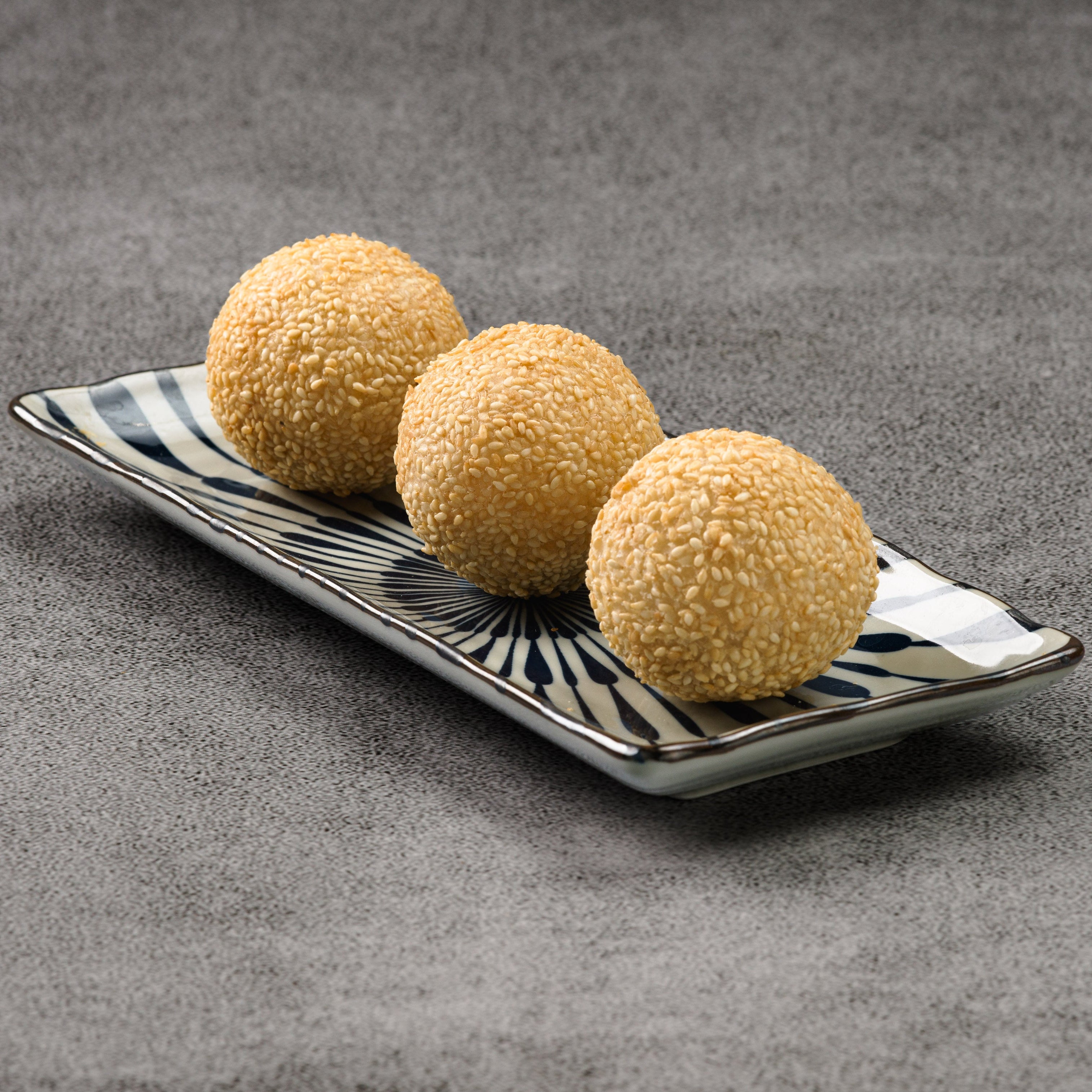 Three sesame balls on a rectangular ceramic plate with a checkered pattern on a gray surface.