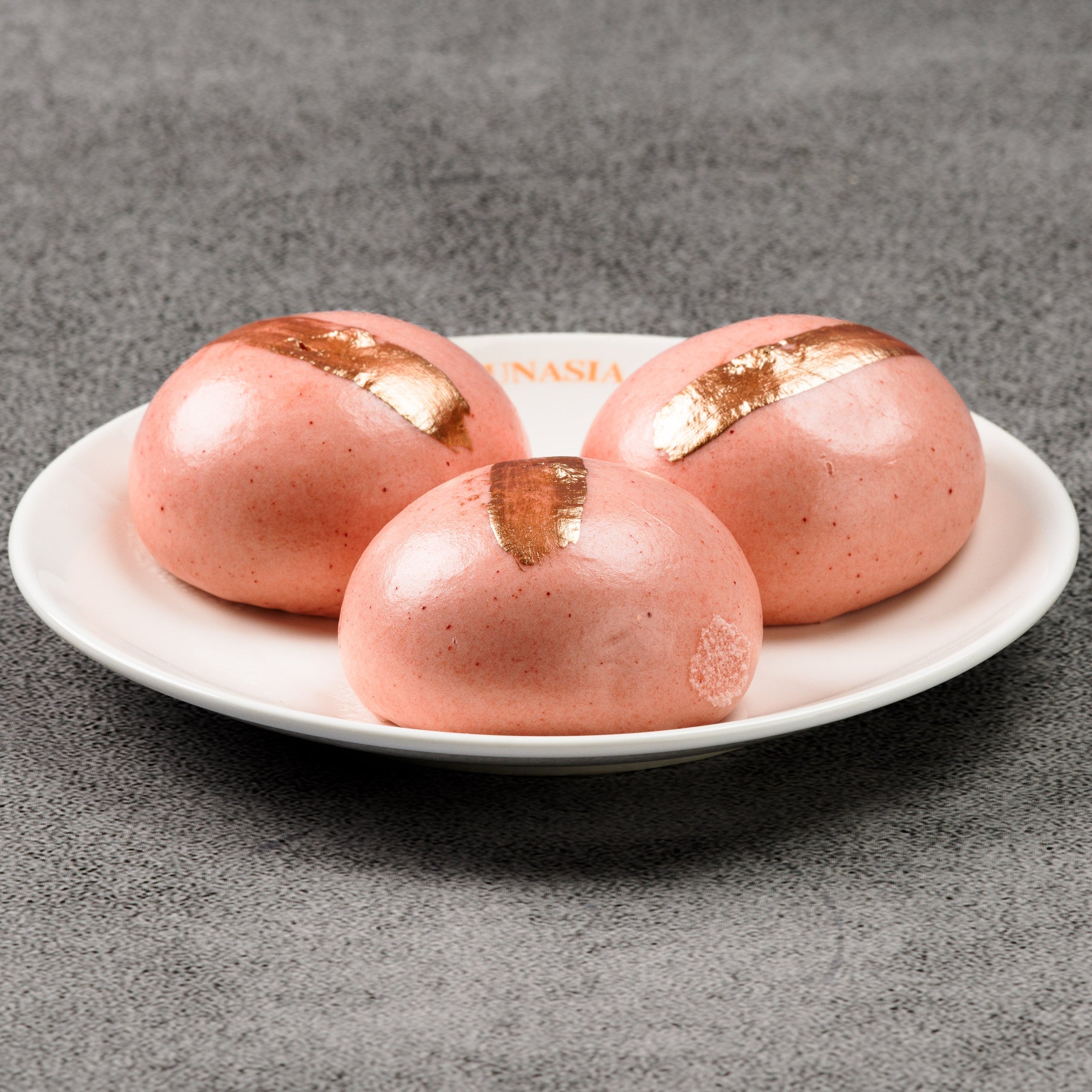 Three pink egg yolk buns with gold accents on a white plate against a gray background