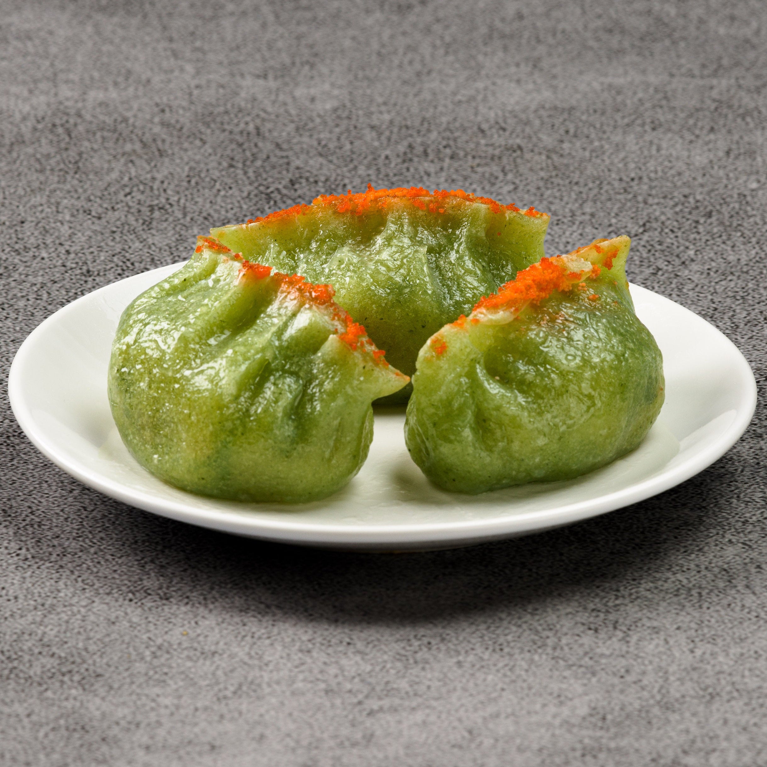 Three green dumplings with orange toppings on a white plate against a gray background