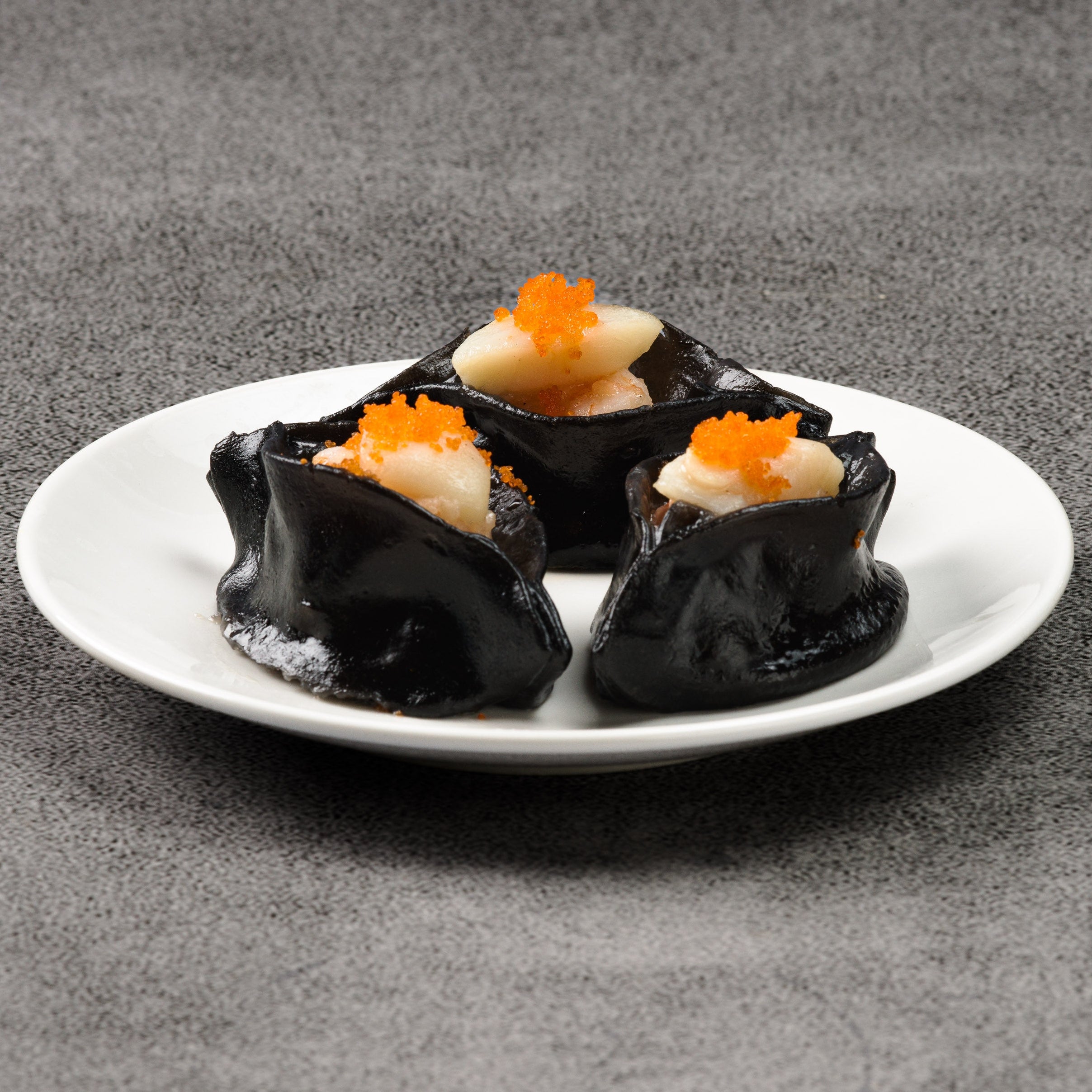 Three black dumplings with orange toppings on a white plate against a gray background